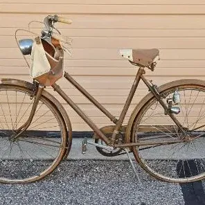 Vintage step-through cruiser bicycle featuring a brown frame, front headlight, handlebar-mounted canvas bag, and rear-wheel generator light.