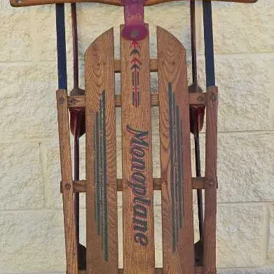 Vintage Monoplane wooden snow sled with metal runners and steering bar, showing original painted graphics and moderate wear.