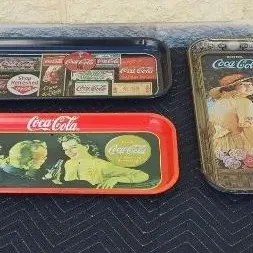 Three vintage Coca-Cola metal serving trays featuring classic advertising graphics and illustrations, showing some light wear and age.