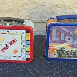 Two vintage-style metal lunchboxes, one featuring a Monopoly board game design and the other featuring Lionel Trains graphics.