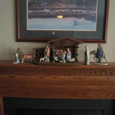 Vintage ceramic nativity set with a wooden stable and additional figurines, displayed on a wooden fireplace mantel.