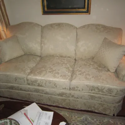 Traditional camelback sofa upholstered in a beige floral damask fabric, featuring rolled arms, a skirted base, and two matching throw...