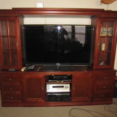 Large cherry wood entertainment center with glass-front side cabinets, lower storage drawers, and a central TV bridge, shown housing a...
