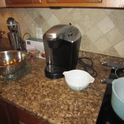 Keurig single-serve coffee maker in black and silver, displayed on a kitchen counter alongside various cookware including a stainless...