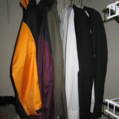 Sale Photo Thumbnail #102: Assorted men's outerwear and formalwear hanging in a closet, including an orange and black active jacket, a tweed blazer, and black suit jackets.