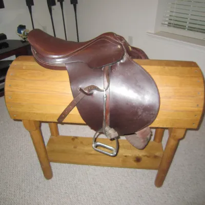 Vintage brown leather English riding saddle with stirrups, displayed on a wooden saddle stand.