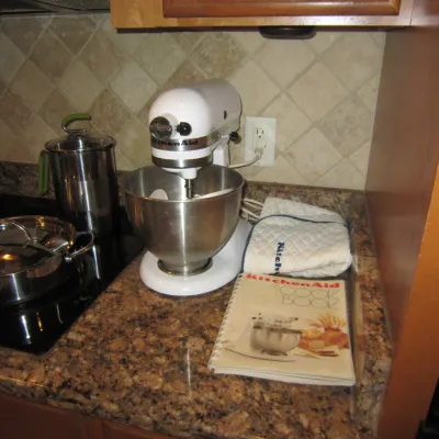 White KitchenAid stand mixer with a stainless steel bowl, accompanied by a KitchenAid cookbook and oven mitt on a granite countertop.
