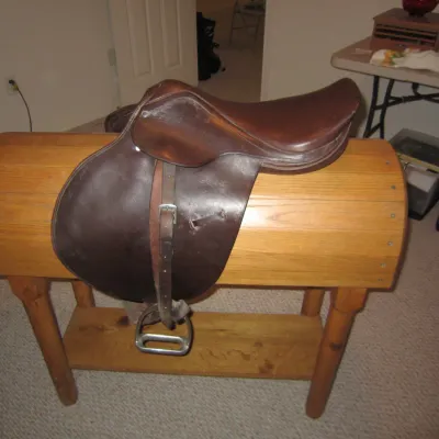 Vintage brown leather English riding saddle with stirrups, displayed on a custom wooden saddle stand.