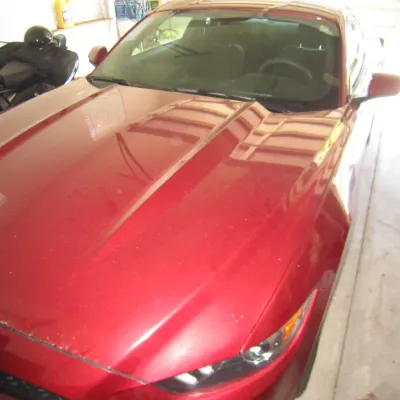Modern red Ford Mustang coupe parked in a garage, showing the hood, windshield, and driver's side headlight.