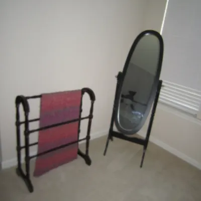 Dark wood cheval floor mirror with an oval frame alongside a matching dark wood quilt rack holding a pink and purple textile.