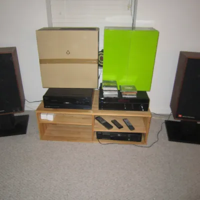 Vintage JBL L26 Decade speakers on black stands, flanking a light wood media console with stereo components, remotes, and original boxes.