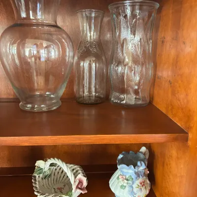 Assorted glassware and decorative ceramics displayed on wooden shelves, including three clear glass vases of varying shapes and two small...