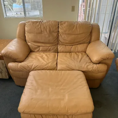 Tan leather loveseat and matching ottoman, showing some wear on the cushions and armrests, available for a buy it now price of $100.