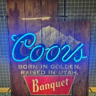 Coors Banquet neon sign shaped like the state of Utah, featuring a faux-wood grain background and the slogan 'Born in Golden. Raised...