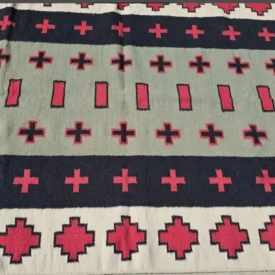 Southwestern-style woven rug featuring a geometric pattern of crosses and stepped motifs in red, black, grey, and cream.