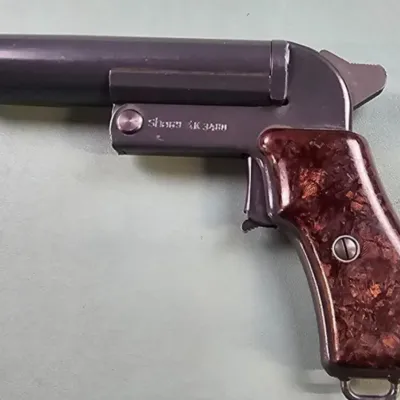 Vintage flare gun featuring a dark metal finish, textured reddish-brown composite grips, and a lanyard loop on the butt.