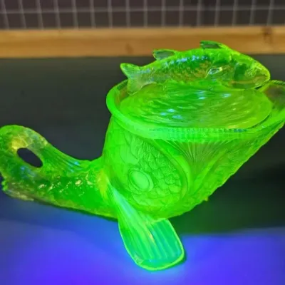 Vintage green uranium glass lidded dish shaped like a fish, glowing brightly under a UV blacklight.