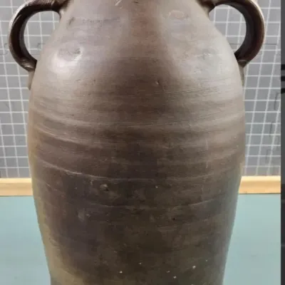 Antique double-handled stoneware jug with a dark brown glaze and a wooden stopper, showing rustic wear and prominent throwing ridges.