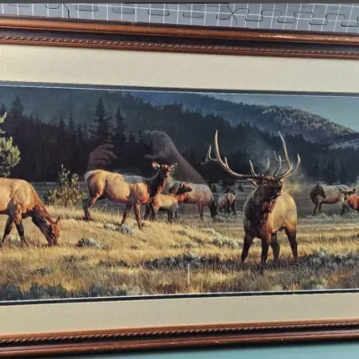 Framed wildlife print depicting a herd of elk in a mountainous landscape, housed in a dark wood frame with a gold inner border.