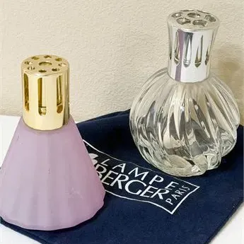 Sale Photo Thumbnail #64: Two Lampe Berger catalytic fragrance lamps, including a frosted lavender conical bottle with a gold-tone cap and a clear swirl-glass bottle with a silver-tone cap, displayed with a blue branded storage pouch.
