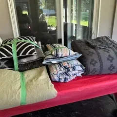 Sale Photo Thumbnail #58: Assorted bedding sets including a grey patterned comforter, a light green quilted coverlet, and striped pillows, displayed on a red table.
