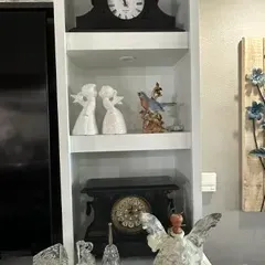 Sale Photo Thumbnail #51: Assorted home decor items displayed on white shelving, including two vintage-style mantel clocks, porcelain angel figurines, a bird figurine, and various glass and metal collectibles.