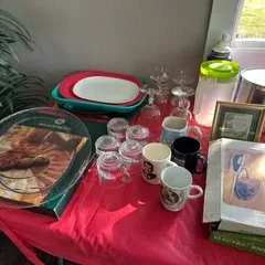 Sale Photo Thumbnail #37: Assorted kitchenware and dining items, including glass platters, mugs, stemware, a plastic pitcher, and a boxed electric knife, displayed on a red tablecloth.
