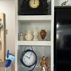 Sale Photo Thumbnail #48: Assorted home decor items displayed on white shelving, including an antique-style black mantel clock, a blue metal table clock, porcelain figurines, and decorative angel statues.