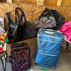 Assorted luggage including a blue rolling suitcase, a black rolling suitcase with a pink lip pattern, and various hanging duffel bags and...