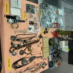 Sale Photo Thumbnail #56: Assortment of costume jewelry, including beaded necklaces, watches, pins, and bracelets, organized on a pink tablecloth with handwritten labels.