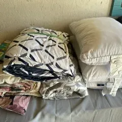 Assorted bedding including folded comforters with geometric and floral patterns, alongside a stack of white bed pillows.
