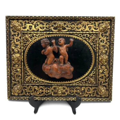 Sale Photo Thumbnail #59: Antique relief plaque featuring two carved wooden cherubs on a cloud, set against a dark background within an ornate, gilded metal frame with cherub head motifs.