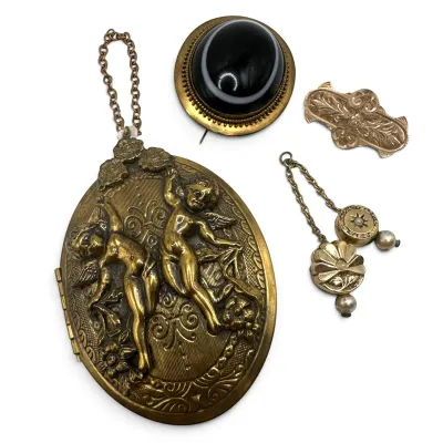 Sale Photo Thumbnail #103: Collection of antique gold-tone jewelry, featuring a large oval locket with embossed cherubs, a black onyx cabochon brooch, an engraved bar pin, and a delicate chain with decorative drops.