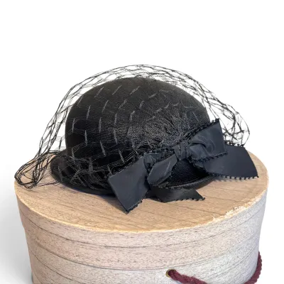 Sale Photo Thumbnail #166: Vintage mid-century black straw tilt hat featuring a birdcage veil and a large satin bow, displayed on a round hat box.