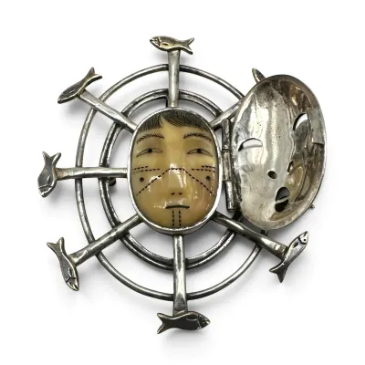 Sale Photo Thumbnail #145: Vintage Alaskan Inuit sterling silver and carved ivory or bone mask brooch, featuring a hinged silver mask cover and a surrounding fish wheel design.