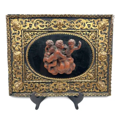 Sale Photo Thumbnail #58: Antique relief plaque featuring carved wooden cherubs on a black background, set within an ornate, gilded metal frame with cherub face motifs.