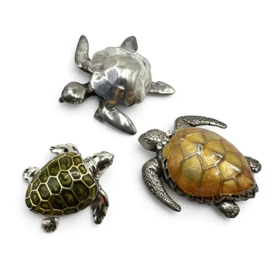 Sale Photo Thumbnail #106: Trio of decorative turtle figurines, including a polished silver-tone metal turtle, a green enameled turtle with silver-tone trim, and a larger yellow-orange enameled turtle with rhinestone collar details.