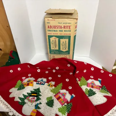 Sale Photo Thumbnail #65: Vintage red felt Christmas tree skirt with sequined snowman and village scene applique, shown alongside a vintage Adjusta-Rite Christmas tree holder in its original box.