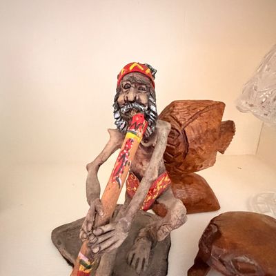 Sale Photo Thumbnail #14: Handcrafted clay figurine depicting an Indigenous Australian man playing a didgeridoo, displayed alongside carved wooden animal sculptures.