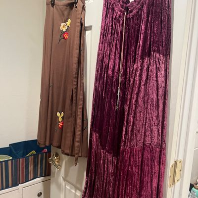 Two skirts hanging on a door, including a brown wrap skirt with embroidered floral details and a long, crushed velvet tier skirt in deep...