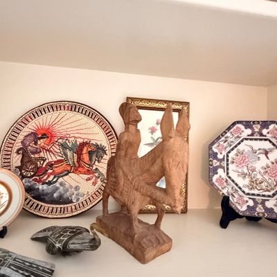 Sale Photo Thumbnail #27: A collection of decorative items on a shelf, featuring a hand-carved wooden sculpture of a figure riding a donkey, flanked by illustrated ceramic plates and small stone carvings.