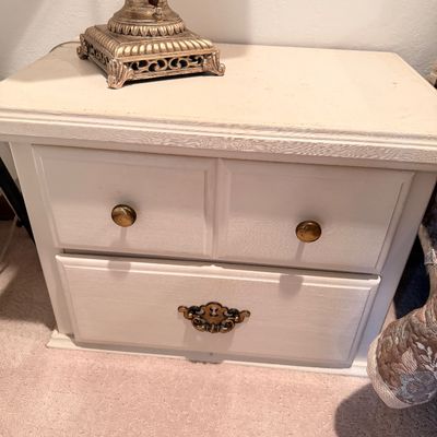 Sale Photo Thumbnail #50: White painted wooden nightstand featuring two drawers with mismatched brass hardware, topped with an ornate gold-finished lamp base.