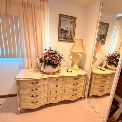 Sale Photo Thumbnail #48: French Provincial style triple dresser in an off-white finish with ornate brass pulls, decorated with a swan planter floral arrangement, a classical figurine, and a matching table lamp.