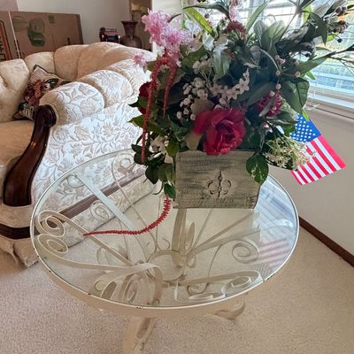 Sale Photo Thumbnail #20: Round glass-top coffee table with a white-painted wrought iron scrollwork base, displaying a faux floral arrangement in a fleur-de-lis planter.