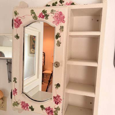 Sale Photo Thumbnail #45: Vintage white-painted wooden medicine cabinet featuring a mirrored door adorned with hand-painted pink floral vine details, alongside open side shelving.