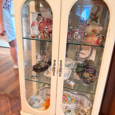 Sale Photo Thumbnail #7: Vintage off-white curio cabinet with arched glass doors, glass shelves, and cabriole legs, filled with various porcelain figurines and decorative dishware.