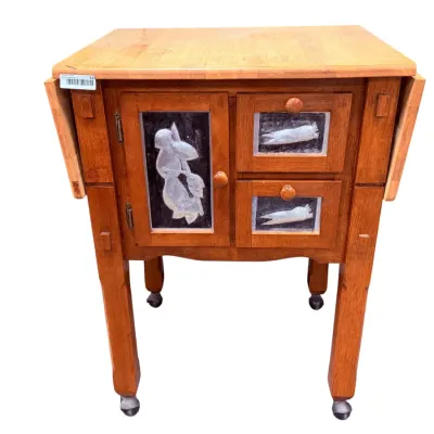Sale Photo Thumbnail #41: Vintage wooden drop-leaf kitchen cart on casters, featuring a butcher block top, one cabinet door, and two drawers with decorative embossed metal panels depicting vegetables.