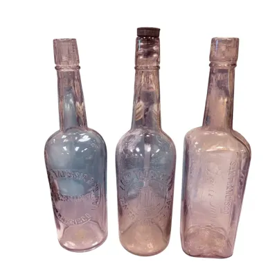 Sale Photo Thumbnail #45: Trio of antique sun-purpled glass bottles from Louis Taussig & Co. of San Francisco, featuring embossed lettering and varying shapes.
