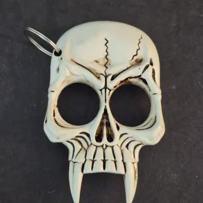 Molded plastic skull-shaped self-defense keychain with large eye holes and pointed fangs, featuring a white finish with dark antiqued...