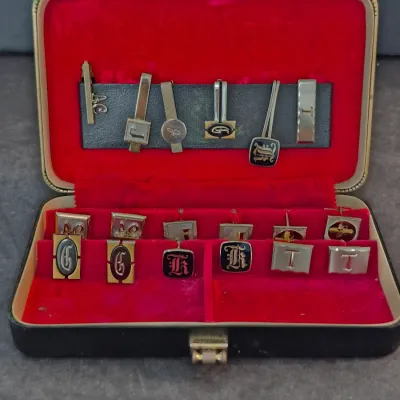 Vintage men's jewelry set featuring assorted monogrammed cufflinks and tie clips, presented in a red velvet-lined display case.